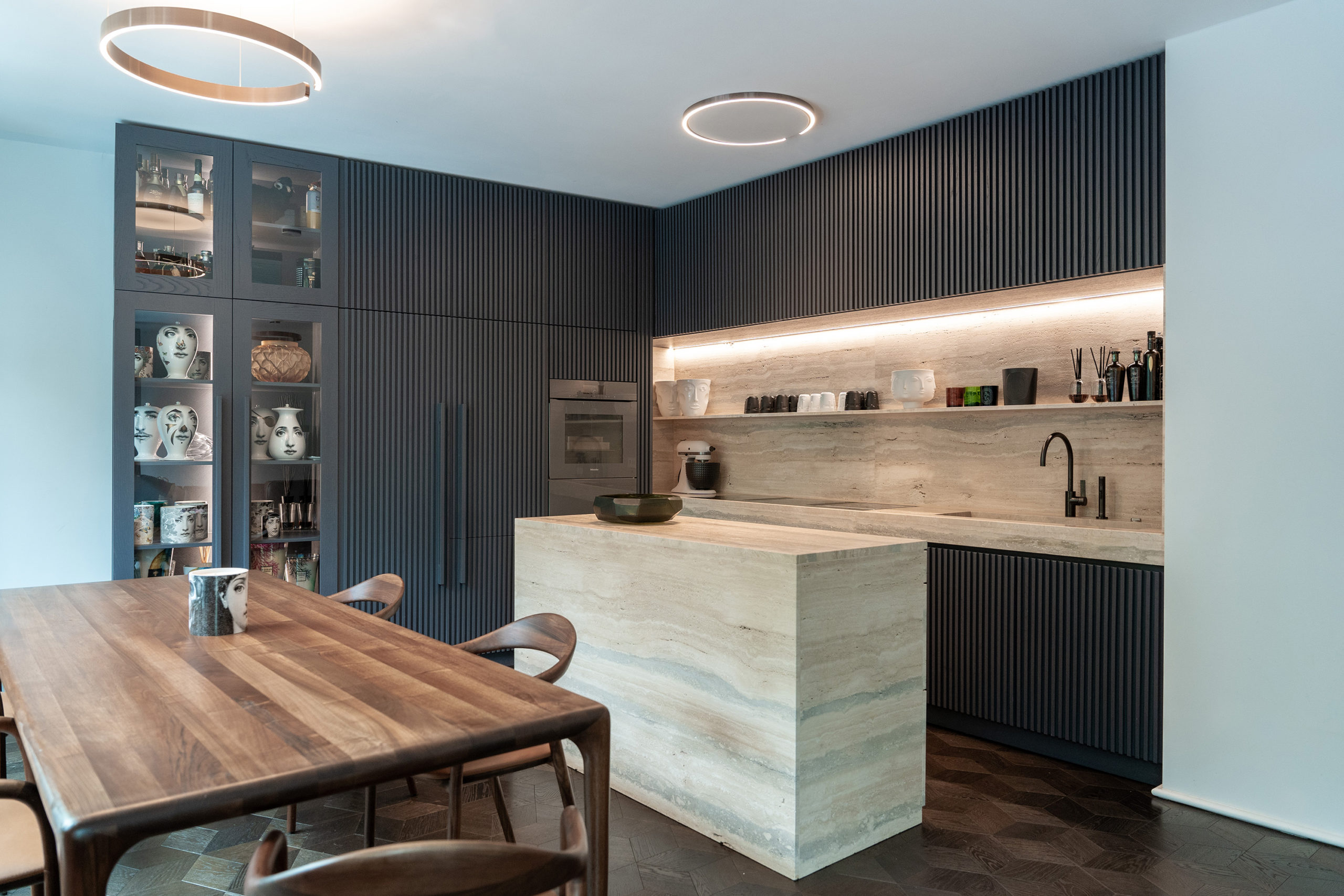 Moderne Designküche mit zentraler Kücheninsel aus Travertin und raumhoher, vertikal strukturierter Holzlamellenwand, hochwertigem Esstisch und skulpturalen Designerstühlen.