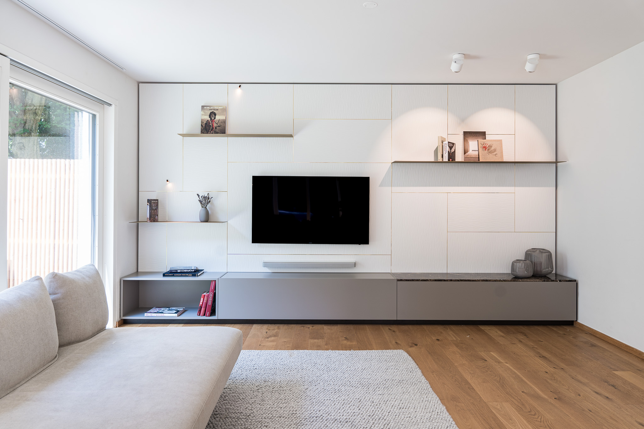Modernes Wohnzimmer mit maßgefertigter Wohnwand in Weiß und Grau, integriertem Fernseher und offenen Regalen – Innenarchitektur München.