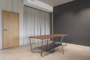 Minimalistischer Besprechungstisch mit Nussbaumplatte und filigranem Metallgestell in einem modernen Büro mit grauer Wandverkleidung, Holzinnentür und Vorhang.